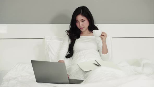 woman drinking a cup of coffee and using a laptop computer on bed