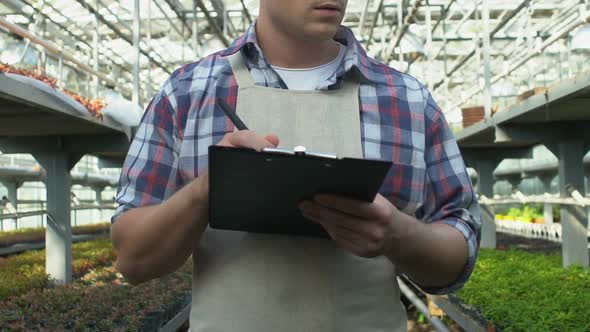 Professional Gardener in Apron Writing Notes on Clipboard Working in Greenhouse