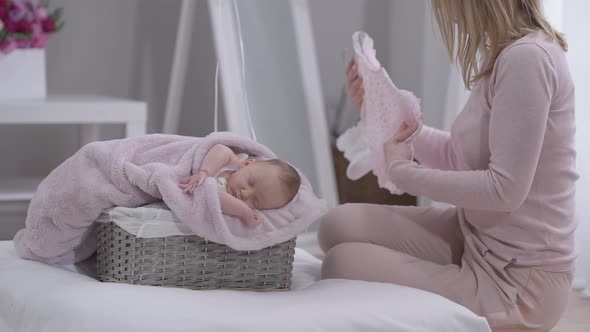 Cute Sleeping Baby Girl in Basket with Unrecognizable Woman Admiring Pink Suit alt