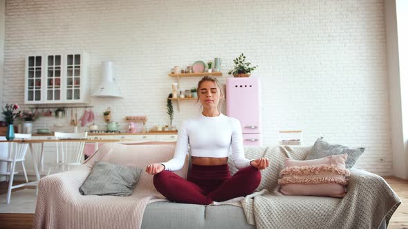 Beautiful Young Woman Meditating on Couch in Lotus Position with Bright Kitchen Background alt