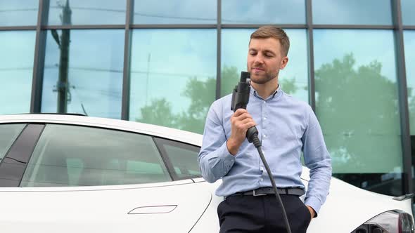 Hansome Guy Standing Near His New Modern Electric Car and Holding Plug of the Charger alt