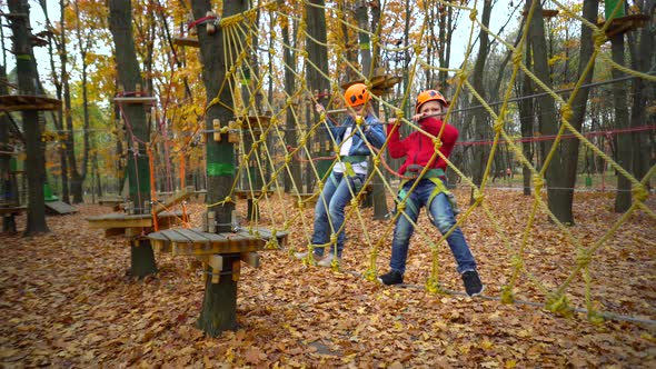 Two little girls in orange helmets and protective gear on rope-way in autumn  forest.