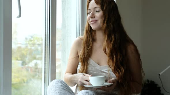 Portrait of woman drinking coffee, sitting on windowsill at window in morning