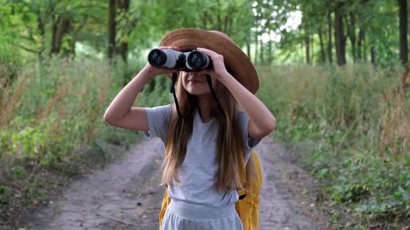 Little Girl Tourist in Hat with Backpack Looking Through Binoculars in the Forest with a Surprised alt
