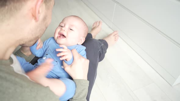 Father Plays with a Smiling Baby Son Lying on Knees at Home Together