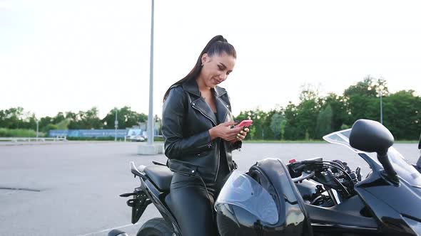 Female Biker Sitting on Black Sporty Motorbike on Mototrack and Using Her Smartphone alt