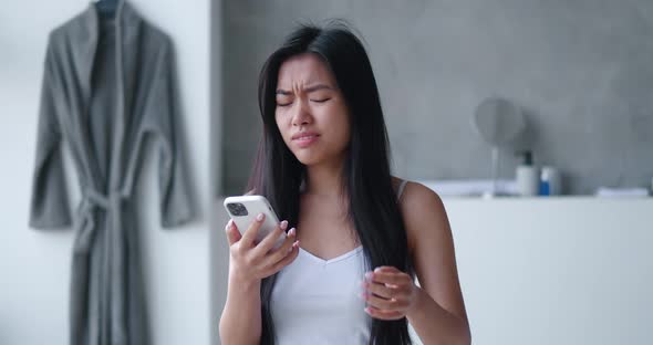 Shocked Asian Woman Reacting on Bad Message on Smartphone Standing in Bathtub alt
