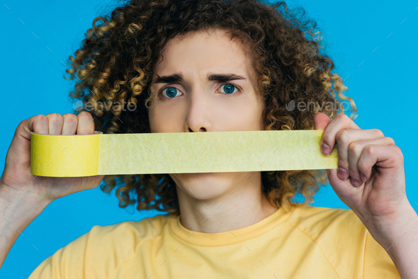 worried curly teenager holding duct tape near mouth isolated on blue