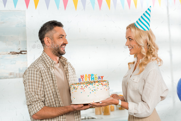 smiling wife presenting birthday cake with happy birthday candles to husband