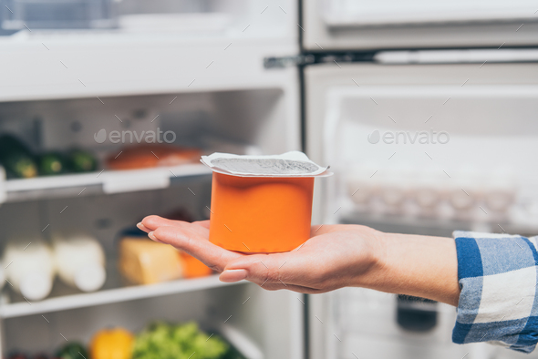 cropped view of woman holding yogurt near open fridge with fresh food on shelves