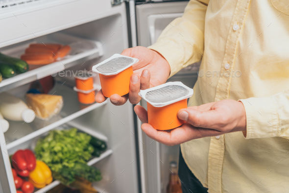 cropped view of man holding yogurt near open fridge full of food