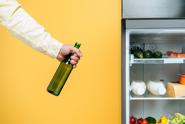 cropped view of man holding yogurt near open fridge with fresh food on shelves isolated on yellow