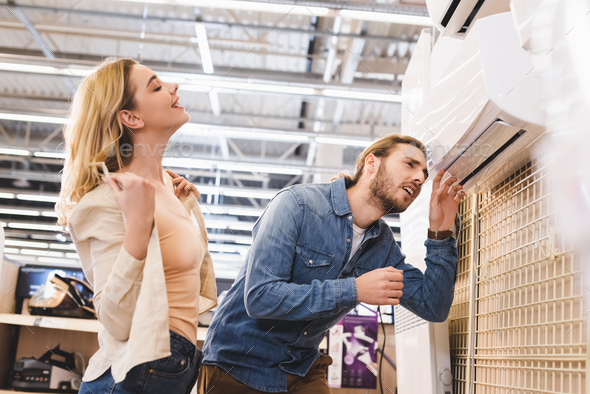 boyfriend looking at air conditioner and smiling girlfriend cooling in ...