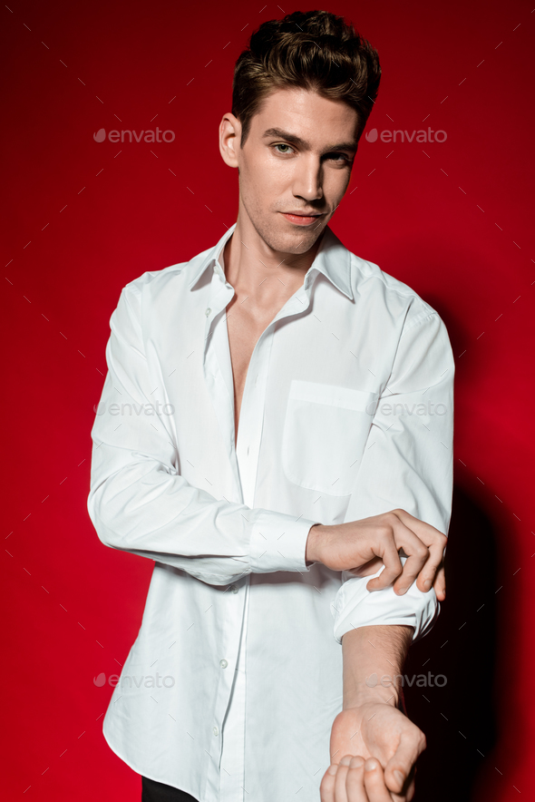 sexy young elegant man in unbuttoned shirt rolling up sleeves on red background