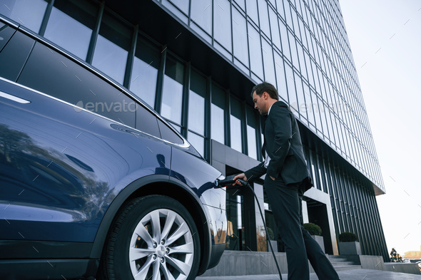 Man in suit and tie is holding charger and standing near his electric car outdoors - Stock Photo - Images