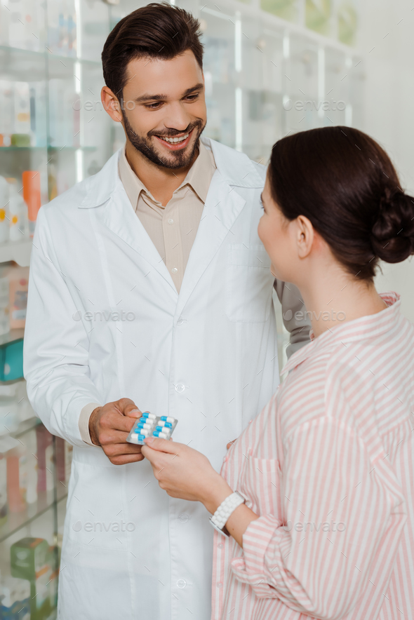 Smiling druggist giving blister with pills to customer by pharmacy showcase - Stock Photo - Images