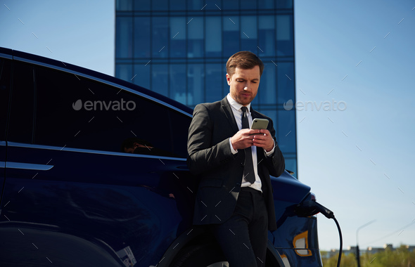 Waiting for a charge. Businessman is standing near his electric car outdoors - Stock Photo - Images