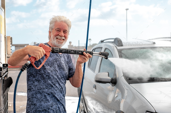 Smiling senior man washing his car in a self-service car wash station using high pressure pump
