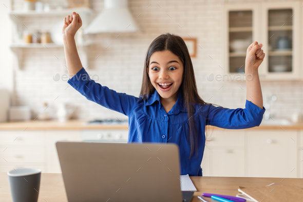 Cheerful Schoolgirl At Laptop Shaking Fists Celebrating News At Home
