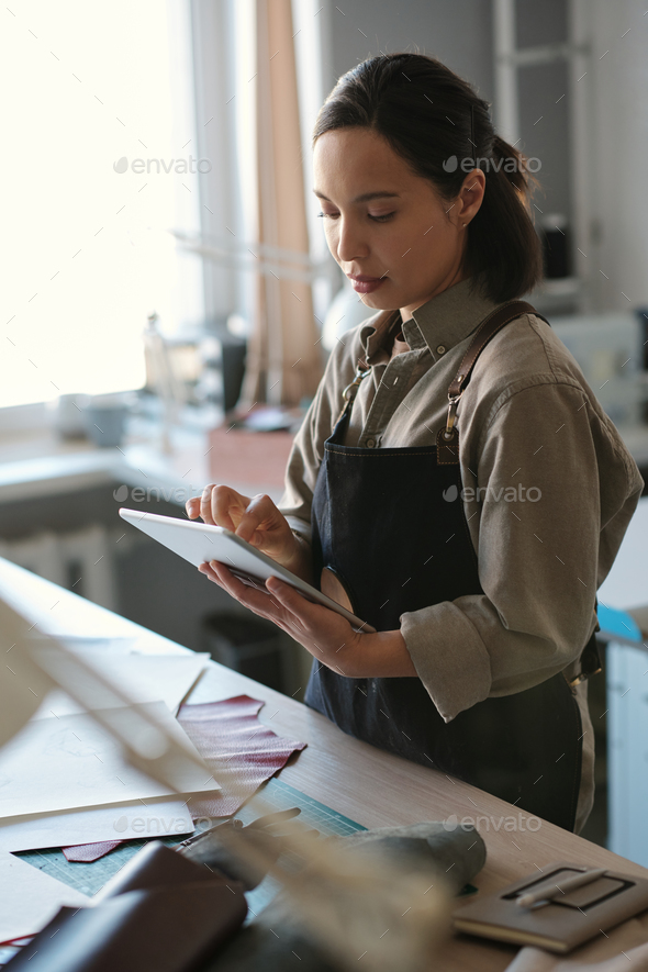 Young serious female artisan in workwear scrolling in tablet by workplace - Stock Photo - Images