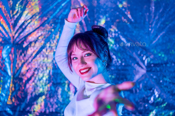 Happy smiling woman in white clothes dancing in neon light and extends a hand inviting you. White