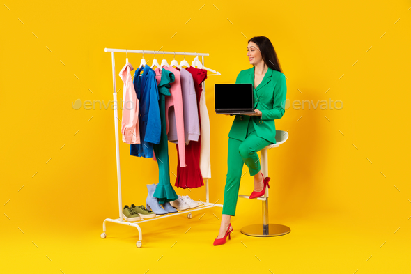 Lady stylist showing laptop with blank screen, advertising store website near clothing rail with - Stock Photo - Images