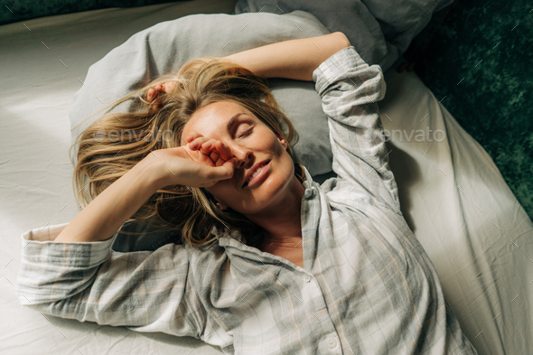 Enjoying serene beautiful woman lies in bed after waking up and stretches and rubs her eyes. - Stock Photo - Images