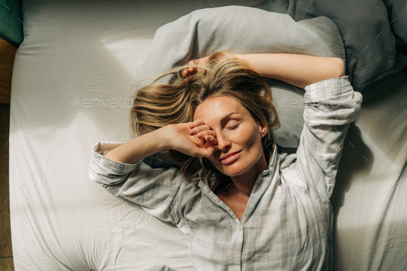 Enjoying serene beautiful woman lies in bed after waking up and stretches and rubs her eyes. - Stock Photo - Images