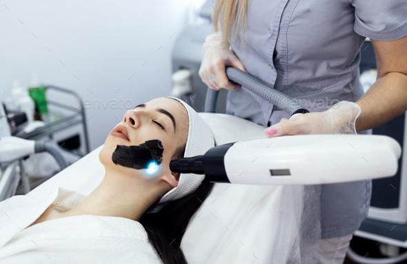Beautician holding beauty laser near woman face covered with carbon mask