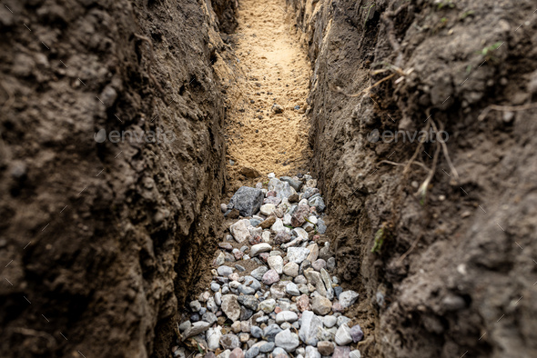 A trench dug in the yard along the fence to lay the drainage pipe,visible gravel and yellow sand. - Stock Photo - Images