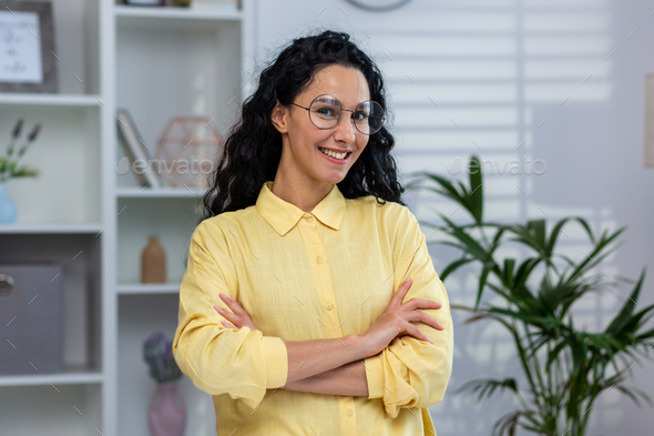 Portrait of young beautiful hispanic at home, woman with curly hair in glasses and yellow shirt - Stock Photo - Images