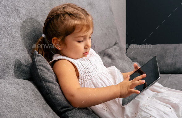 Cute little girl sitting on sofa and using tablet. - Stock Photo - Images