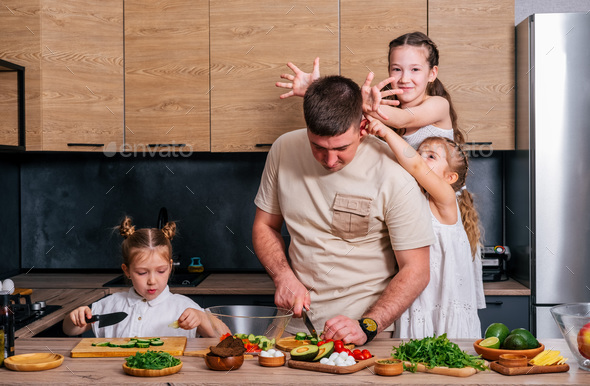 A young father cooks in the kitchen with his daughters and they give him horns and fool around. - Stock Photo - Images