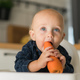 Happy baby sitting in high chair eating carrot in kitchen copy space. Healthy nutrition for kids - PhotoDune Item for Sale