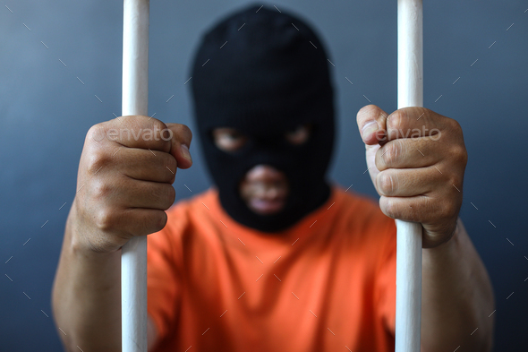 A prisoner in orange shirt and black mask inside the bars of a prison  - Stock Photo - Images