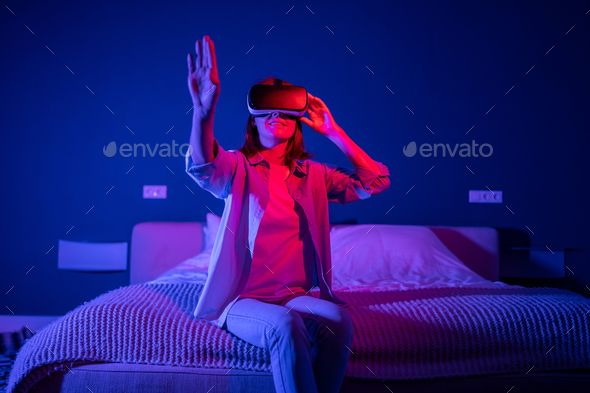 Joyful 3D video game female in VR headset lost touch with reality, sit at bed at home, neon light - Stock Photo - Images