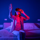 Joyful 3D video game female in VR headset lost touch with reality, sit at bed at home, neon light - PhotoDune Item for Sale