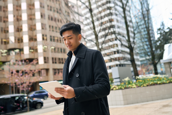 Young smiling Asian business man walking in city using digital tablet. - Stock Photo - Images