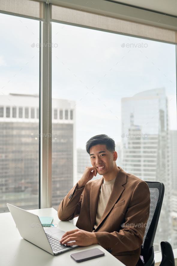 Young happy Asian businessman looking at camera working on laptop in office. - Stock Photo - Images