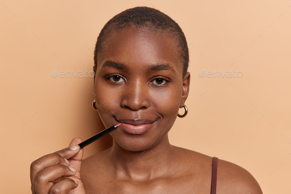 Horizontal shot of short haired black woman holds red lip pencil puts on makeup dressed in casual t