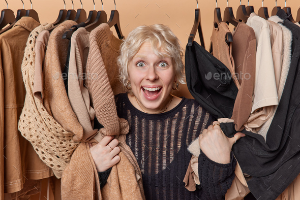 Cheerful surprised young blonde woman stares impressed poses among stylish clothes hanging on rail - Stock Photo - Images