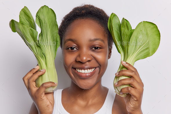 Headshot of positive dark skinned woman holds green bok choy vegetables near head eats healthy food - Stock Photo - Images