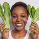 Headshot of positive dark skinned woman holds green bok choy vegetables near head eats healthy food - PhotoDune Item for Sale