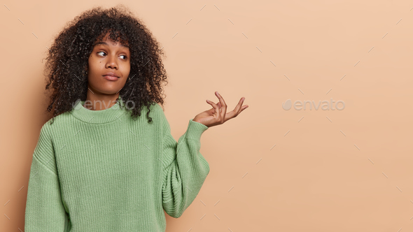 Perplexed woman with dark skin curly hair keeps hands spread sideways reflects sense of confusion
