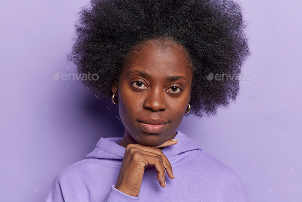 Portrait of dark skinned woman with curly bushy hair keeps hand under chin focused attentively at - Stock Photo - Images