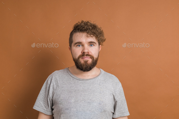 Close up portrait curly European male with little beard wears casual t-shirt over brown background - Stock Photo - Images