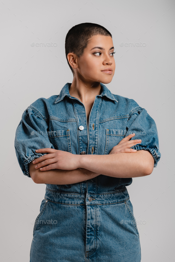 Confident young shaved head woman in denim clothes keeping arms crossed against grey background