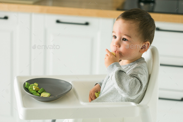 Happy baby sitting in high chair eating fruit in kitchen. Healthy nutrition for kids. Bio carrot as - Stock Photo - Images