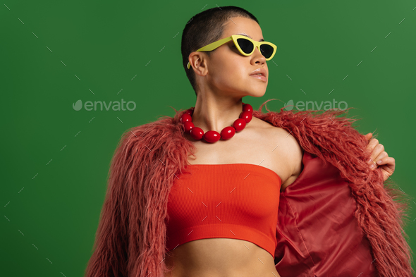 Beautiful short hair woman in trendy glasses adjusting her fluffy jacket against green background - Stock Photo - Images