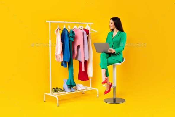 Beautiful woman stylist sitting on yellow background near lot of clothes on garment rack and using
